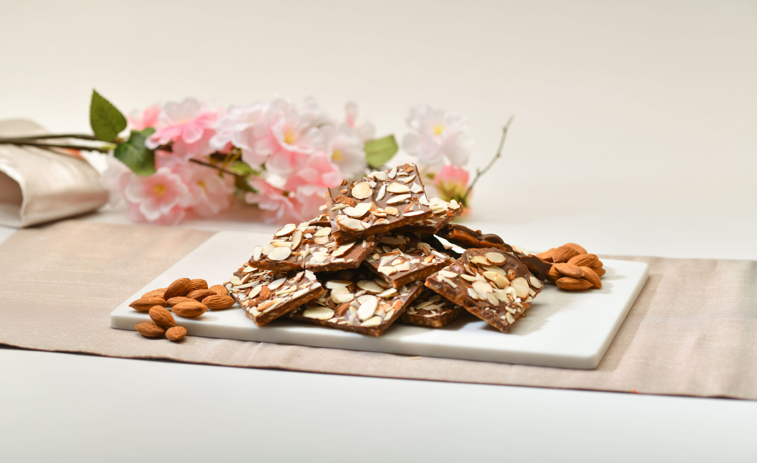 Stacked pieces of Almond Tree handcrafted almond toffee on a marble board, with cherry blossoms behind